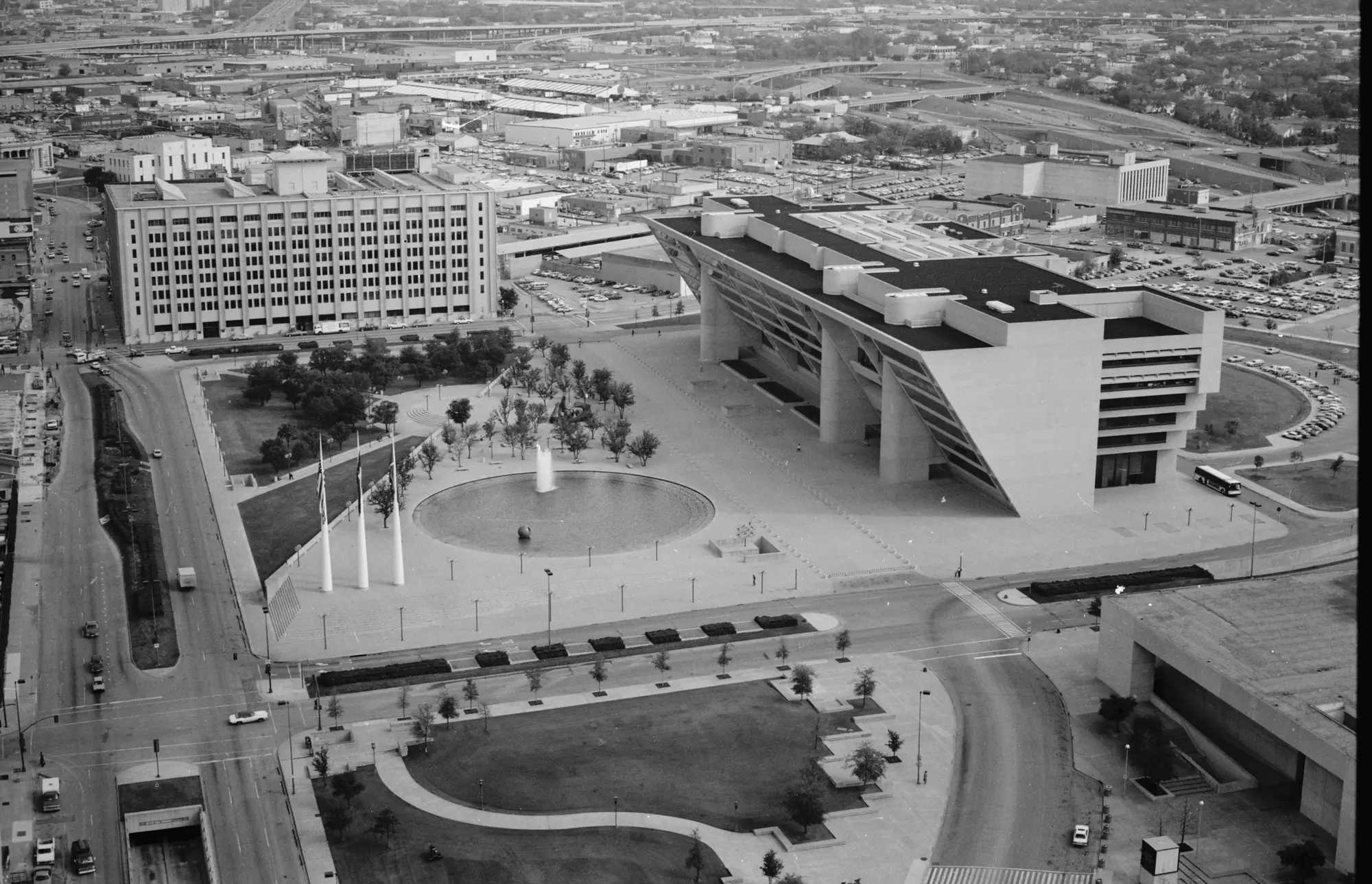 Aerial photograph looking east across City Hall Plaza. Dallas City Hall sits at right, its upper floors cantilevered out over the plaza; a circular fountain, three flagpoles, and rows of young trees fill the open ground in front of it.