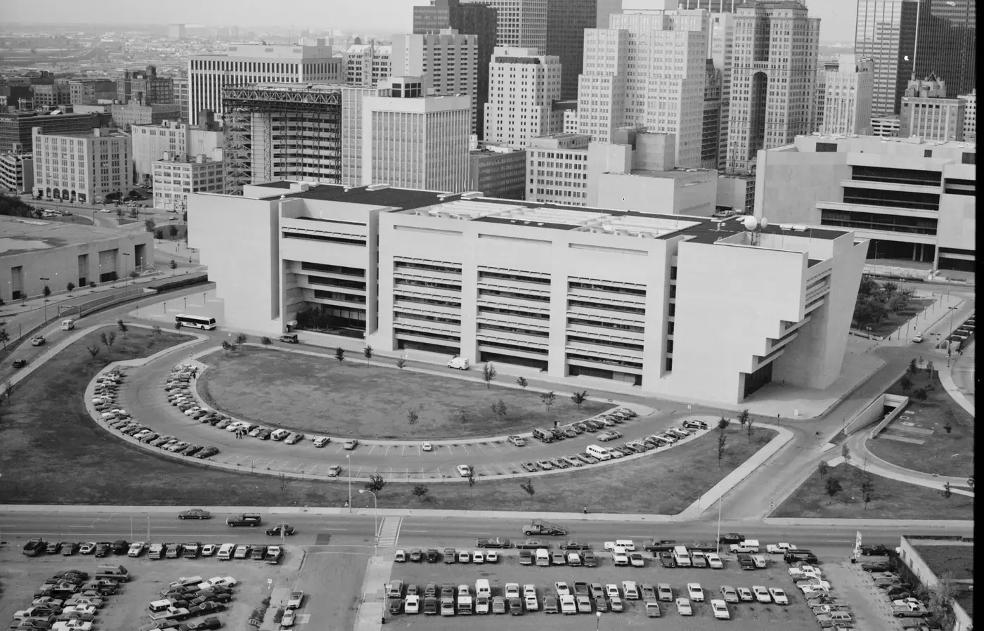 Aerial photograph of the south side of Dallas City Hall, looking north. The downtown skyline rises behind the building; a curved drive, a lawn, and large surface parking lots occupy the foreground.